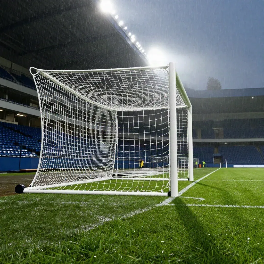 Nasses Fußballtor unter Flutlicht, Nebel und leere Tribüne am Abend.